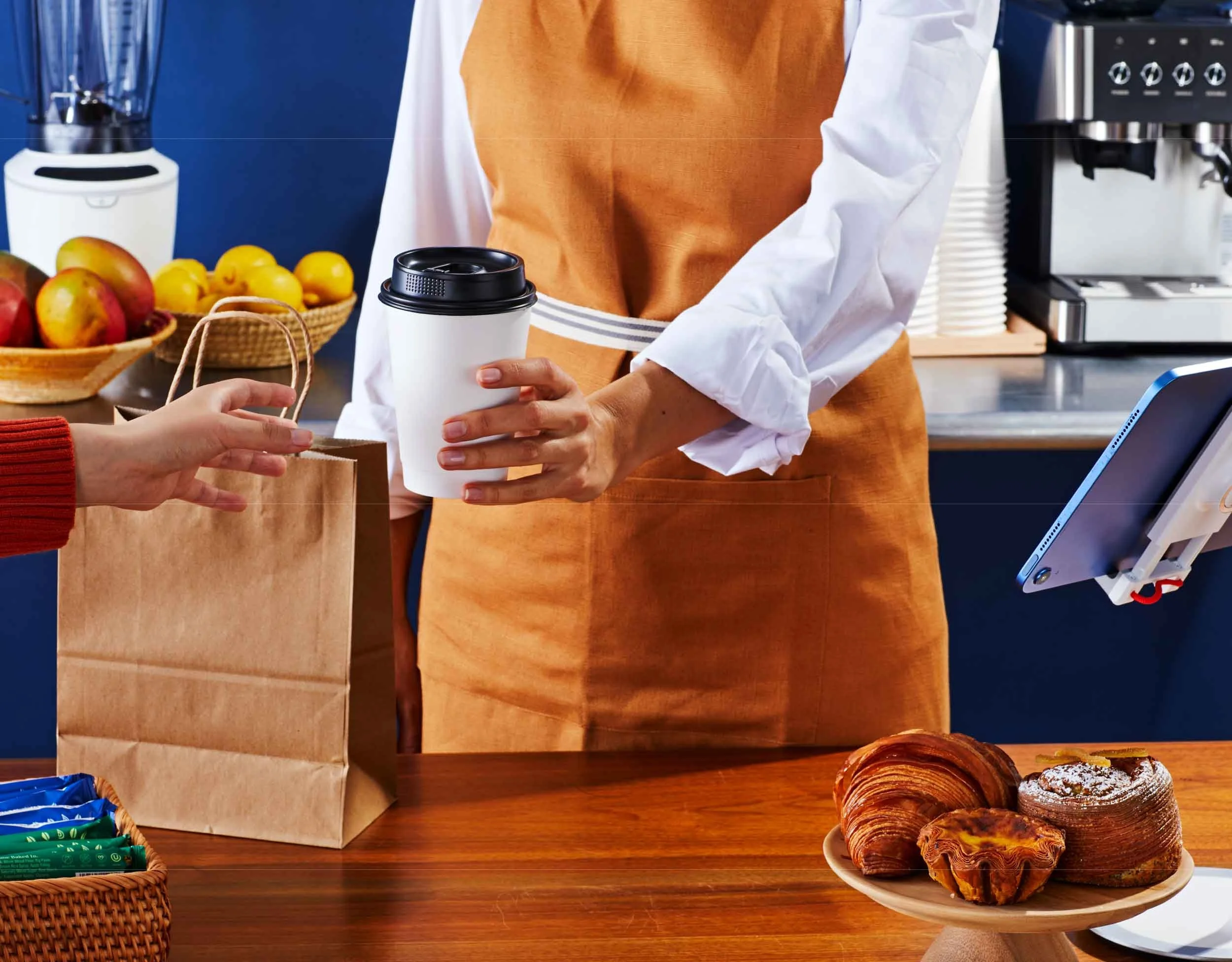 Person handing a paper bag filled with pastries to another person at a breakfast or coffee shop counter, with sliced croissants in a bowl and apples and lemons in baskets on the counter.