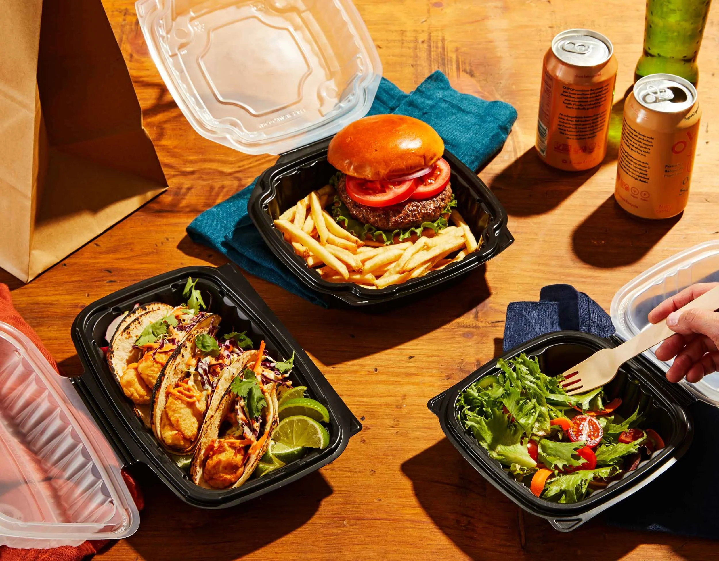 A black container holding a cheeseburger with tomato, lettuce, and onion, served with French fries and lime wedges. Other containers with tacos, salad, and side dishes are on a wooden table, with canned drinks and a green bottle nearby.