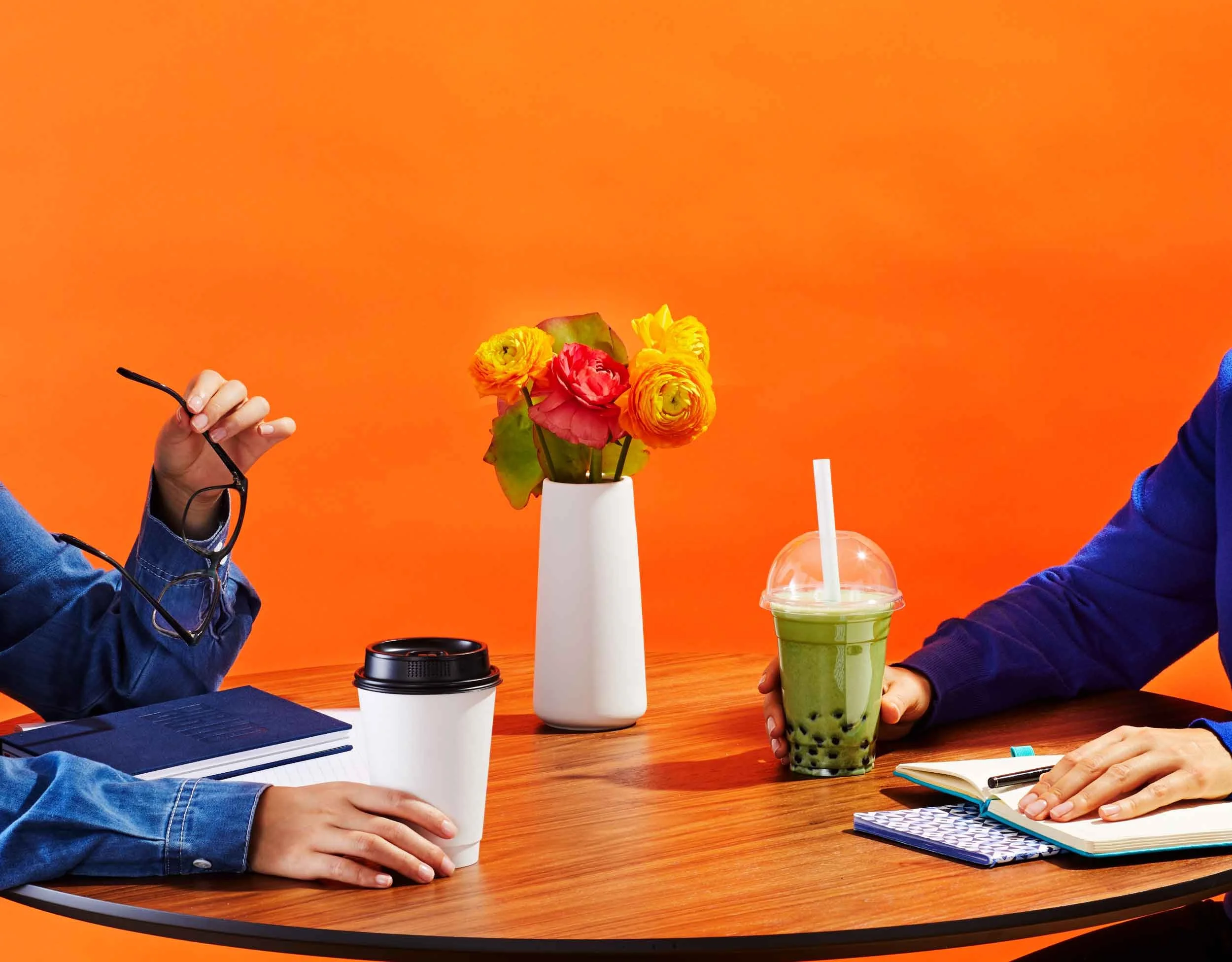 Two people sitting at a wooden table with beverages, a notebook, and a vase with colorful flowers against an orange background.