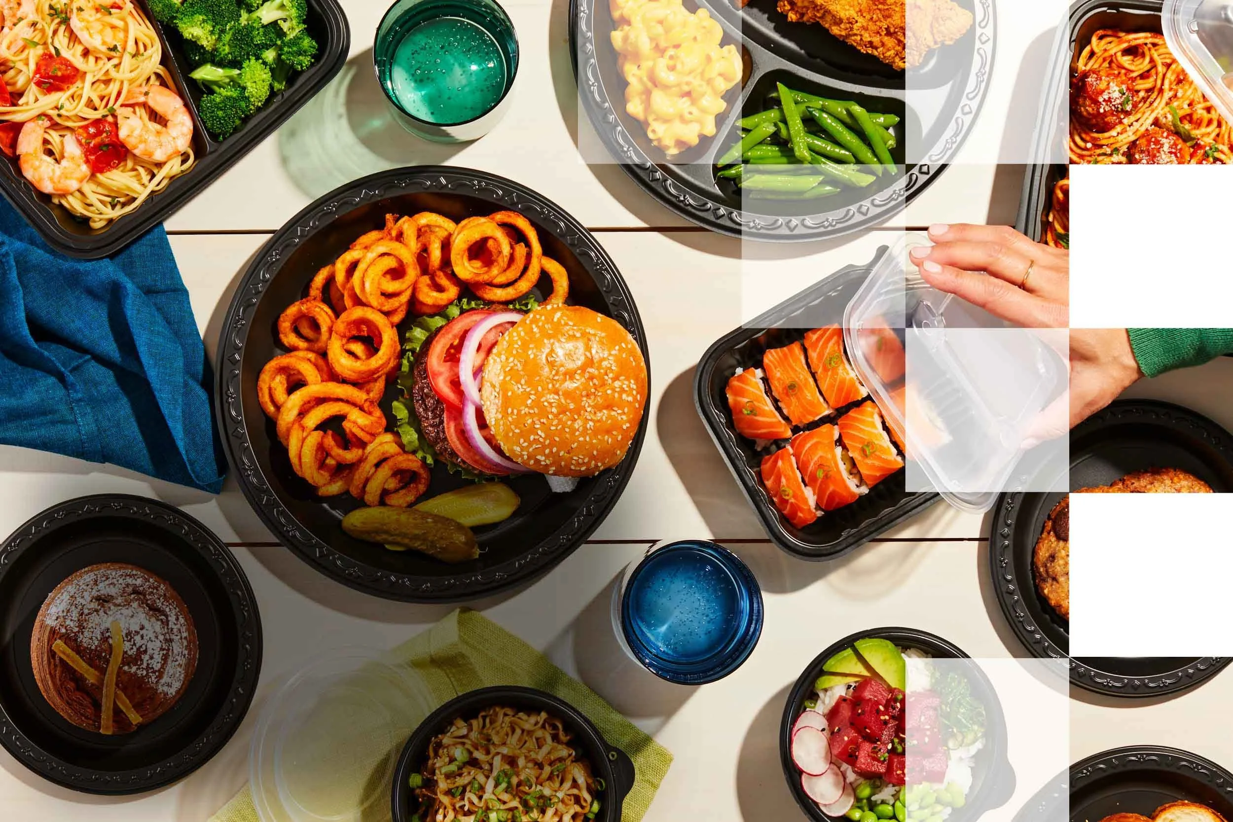 Assorted takeout dishes including pasta with shrimp, broccoli, fried chicken, green beans, sliders with cheese, sushi, salad with radish, watermelon, and grape slices, and a glass of blue drink on a white table.