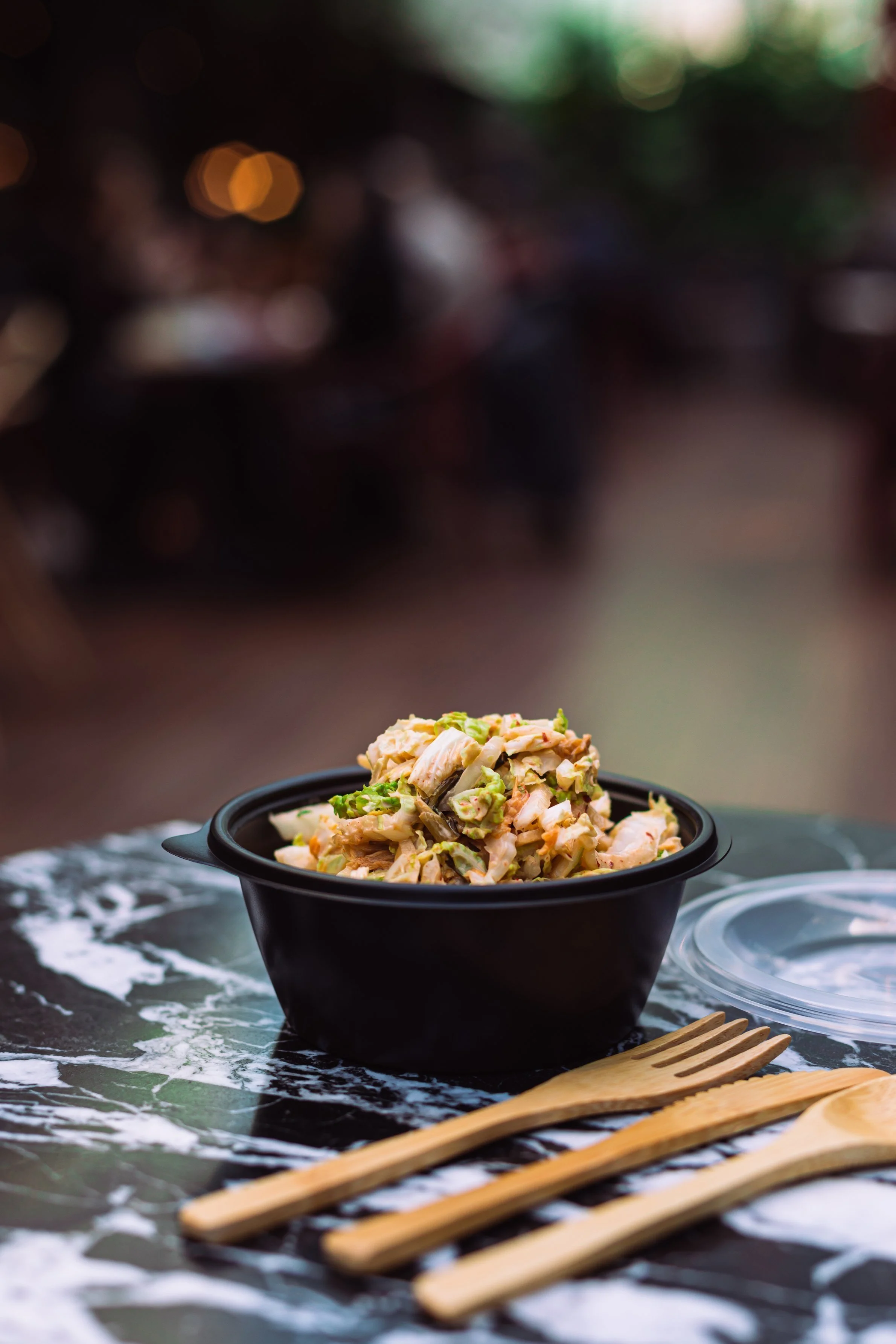 A black bowl filled with chicken salad, placed on a black marble surface. Wooden utensils and a clear plastic lid are nearby. The background is dark and blurred.