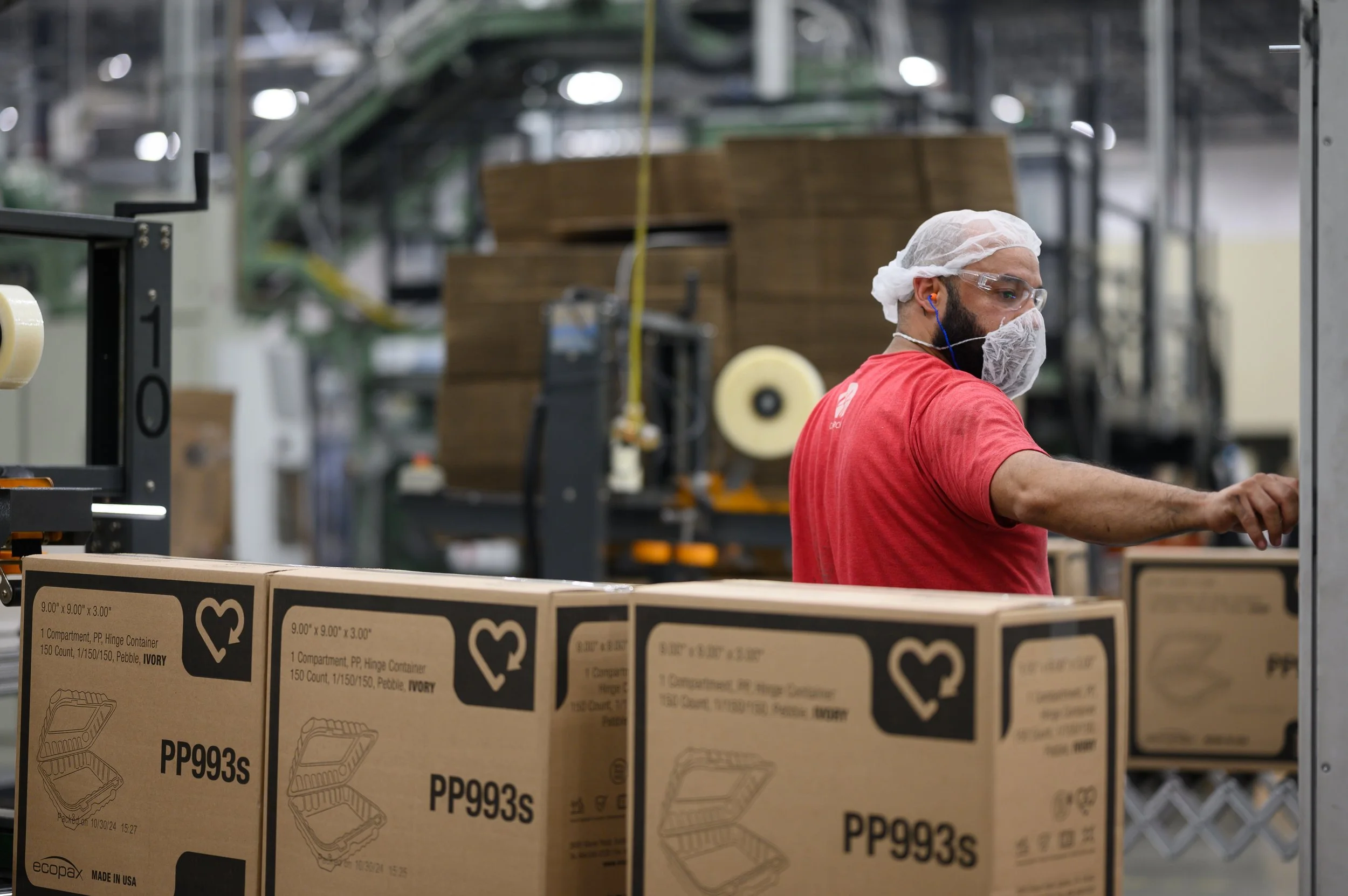 A worker in a factory wearing a hairnet, safety glasses, and a face mask, working on a production line with cardboard boxes labeled PP993S.
