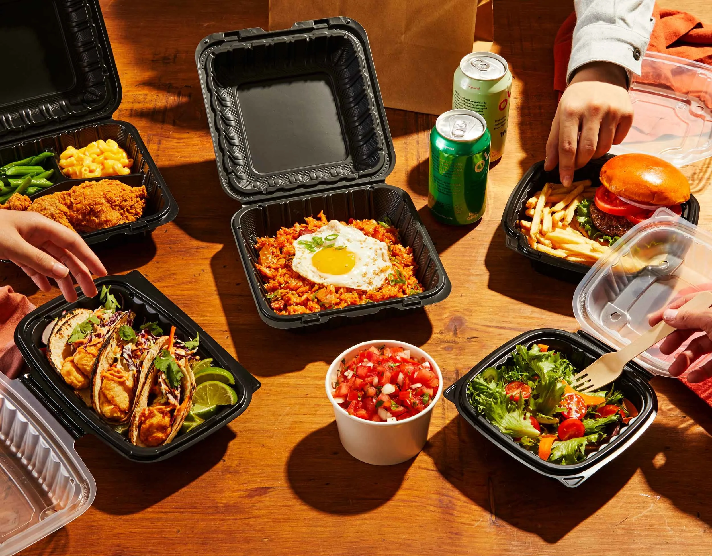 Several takeout food containers on a wooden table with fast food items including salad, tacos, fried chicken, pasta, a cheeseburger with fries, and cans of soda.