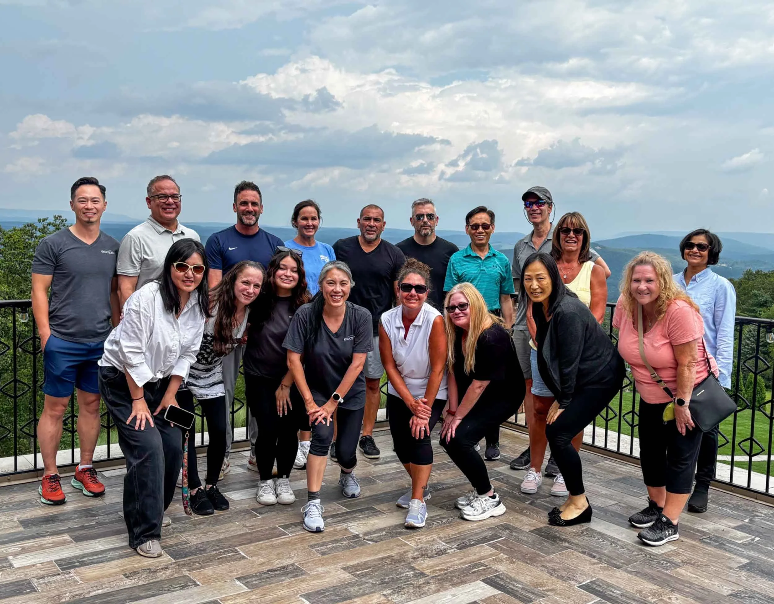 Group of diverse people smiling and posing for a photo outdoors on a wooden deck with scenic mountains and sky in the background.