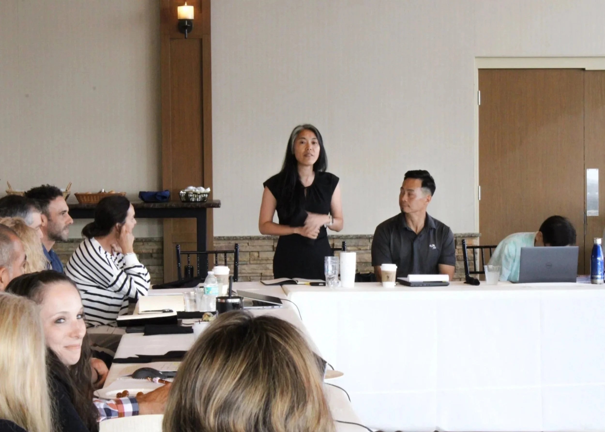 A woman in black is standing and speaking in front of a group of seated people in a conference or meeting room. Several audience members are visible, some taking notes. The room has a beige wall and a wooden door.