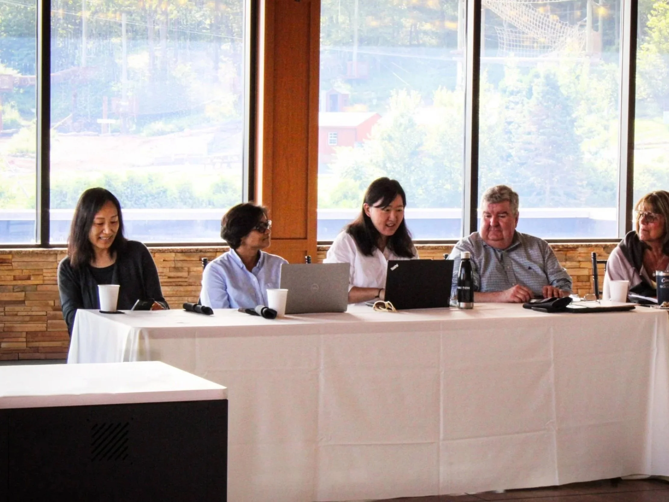 People seated at a long conference table with laptops, coffee cups, and microphones inside a room with large windows showing an outdoor landscape.