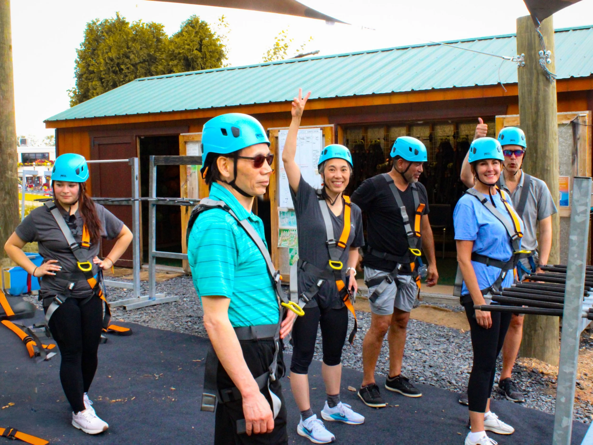 Group of five people wearing blue helmets and safety harnesses standing outdoors in front of a wooden building, some smiling, one person raising two fingers in a peace sign, another giving a thumbs-up.