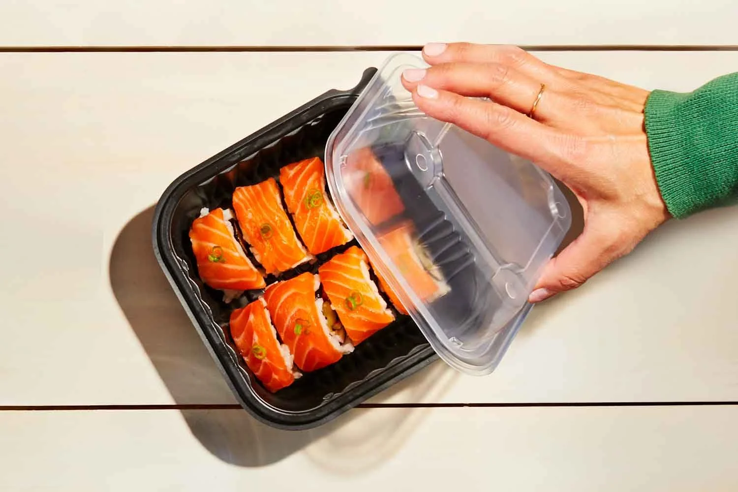 A person's hand opening a black plastic container of sushi rolls with salmon on top, placed on a white table.