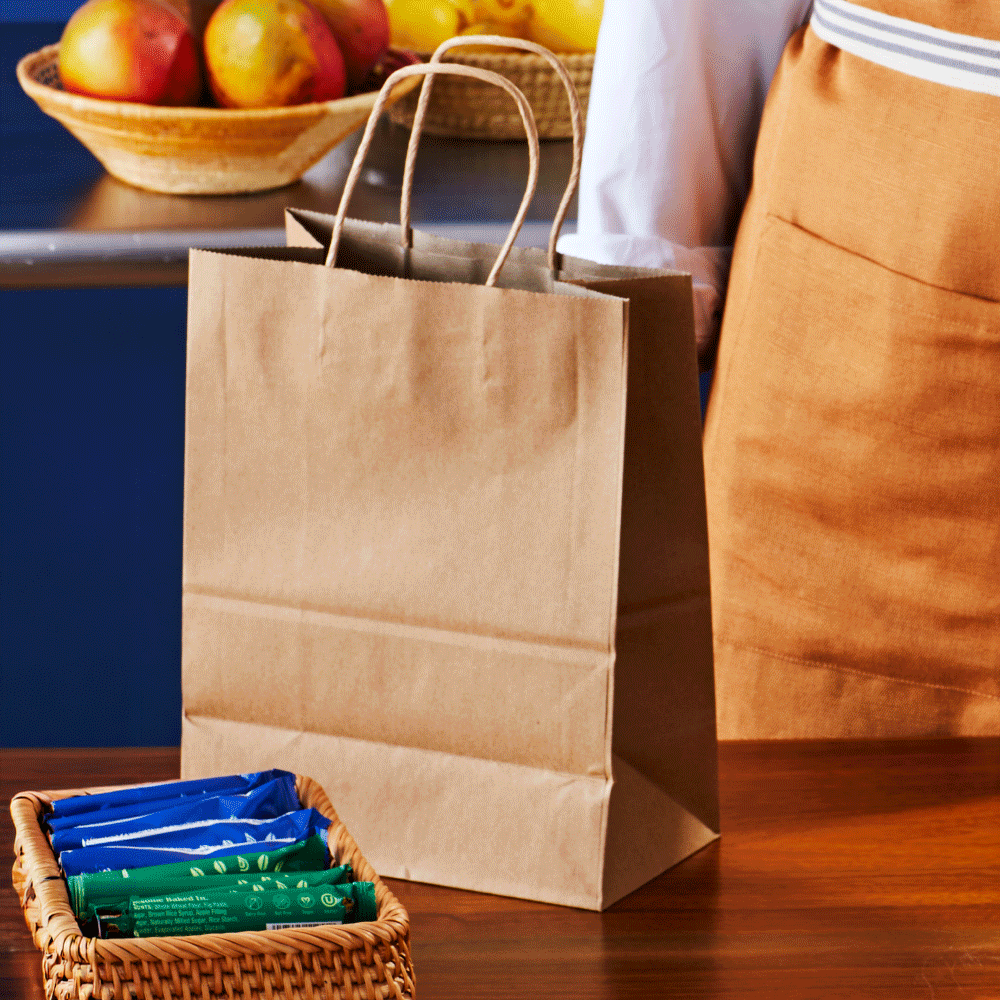 A brown paper shopping bag on a wooden table with colorful snack bars in a woven basket in front of it. In the background, a person wearing an orange apron is standing near a blue counter with a bowl of apples.