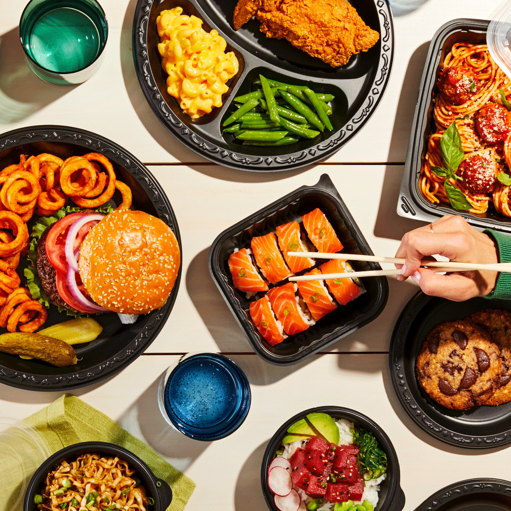 Assorted takeout food on a table including a burger, fried chicken, sushi, spaghetti, salad, cookies, and small bowls of various side dishes, with a woman's hand picking up sushi with chopsticks.