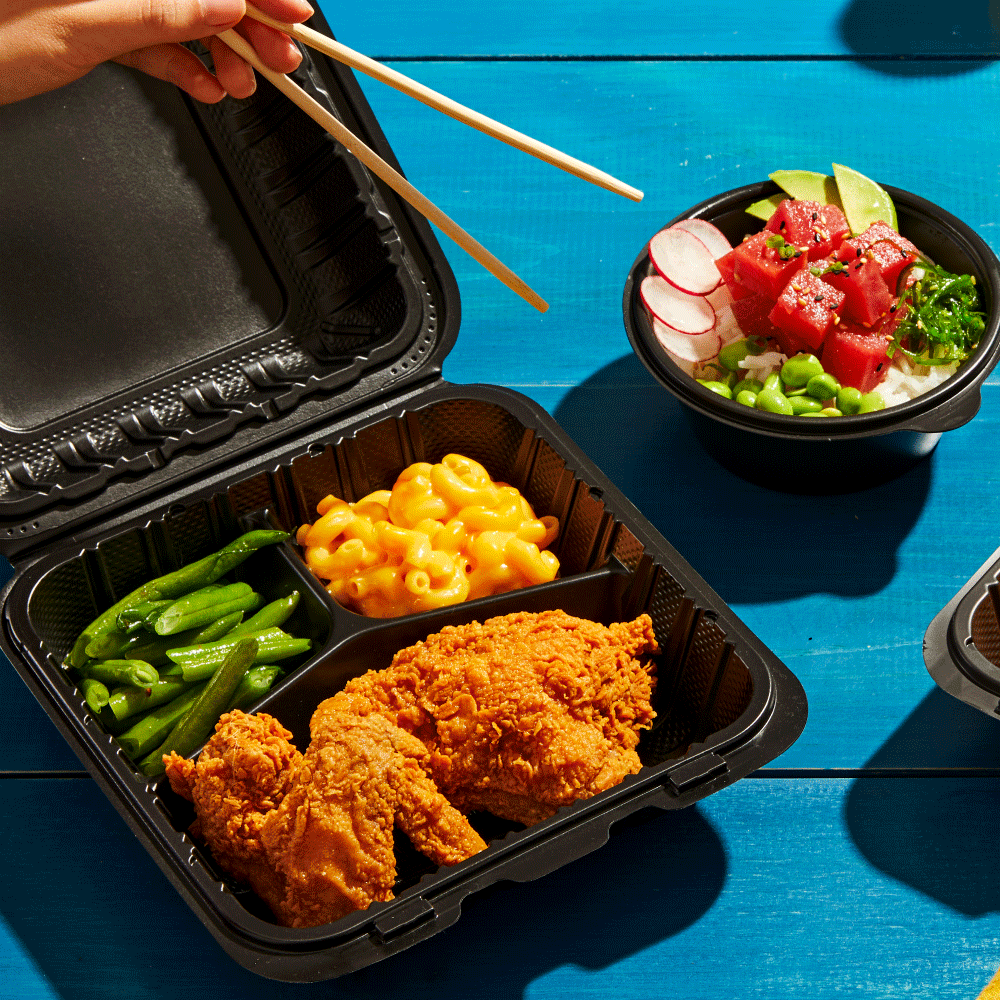 A black bento box with fried chicken, macaroni and cheese, and green beans. A hand holding chopsticks about to pick up food. A black bowl with a salad containing watermelon, radishes, cucumbers, edamame, and greens on a bright blue table.
