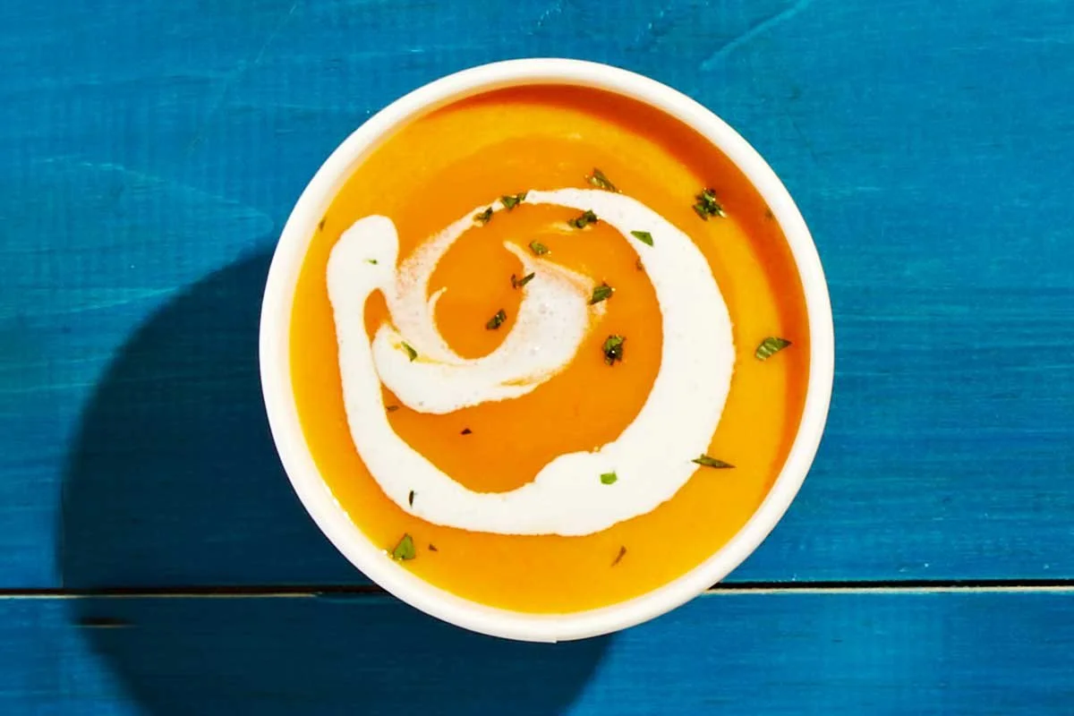 A bowl of creamy orange soup garnished with a swirl of white cream and sprinkled with chopped herbs on a blue wooden surface.