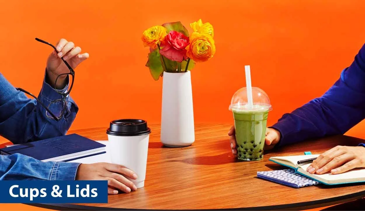 Two people sitting at a table with drinks, a notebook, pen, and a vase of colorful flowers, against an orange wall.