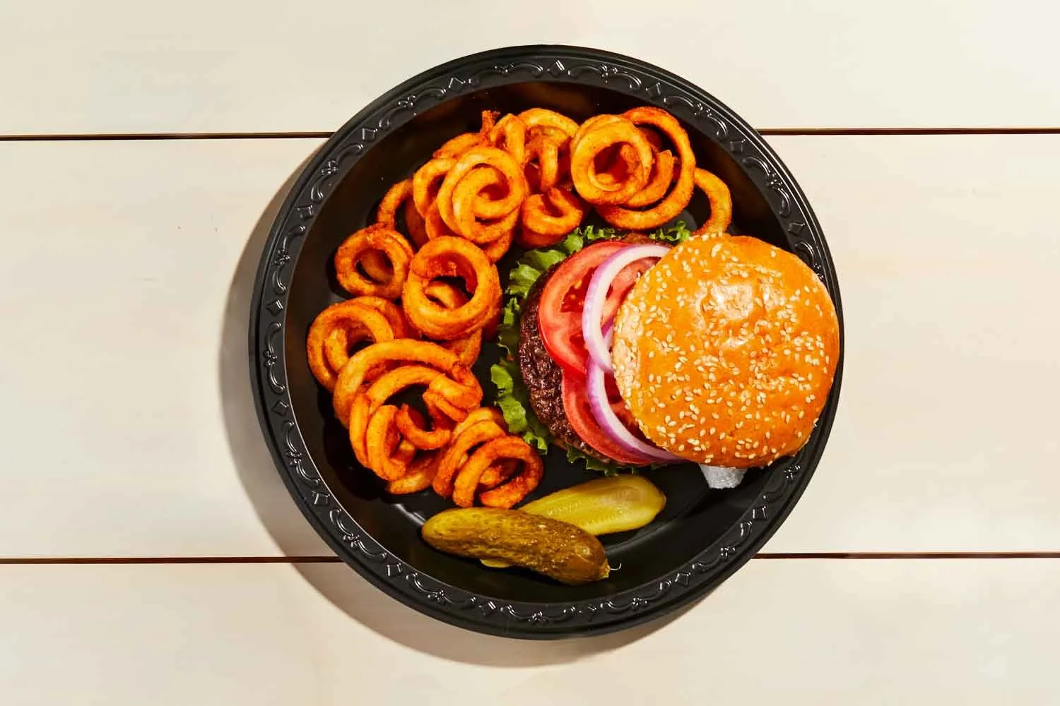 Plate of crispy curly fries, cheeseburger with lettuce, tomato, onions, and sesame seed bun, accompanied by pickle slices.
