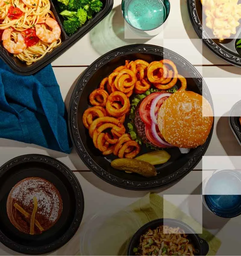 Assorted takeout dishes including pasta with shrimp, broccoli, fried chicken, green beans, sliders with cheese, sushi, salad with radish, watermelon, and grape slices, and a glass of blue drink on a white table.