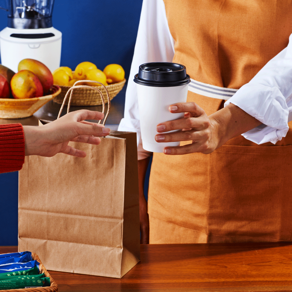 Person handing a paper bag to another person who is holding a coffee cup in a kitchen with apples and lemons in the background.