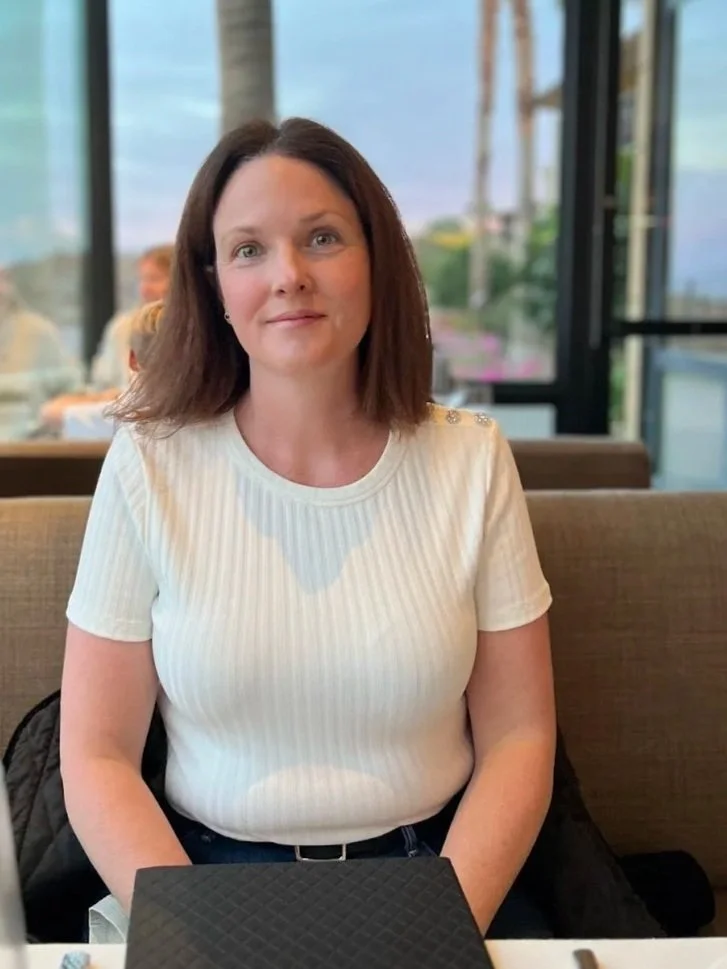 A woman with shoulder-length brown hair sitting at a restaurant table with a large window behind her showing trees and water.