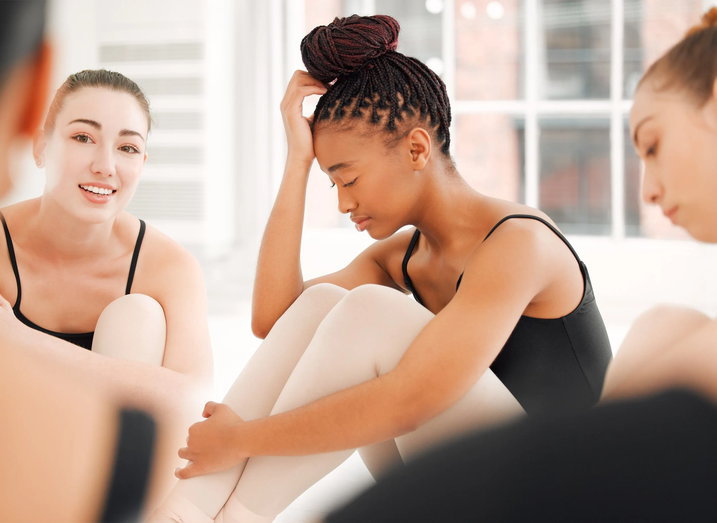 A group of young women in ballet attire sitting in a circle, some smiling, one looking down with a hand on her forehead, in a bright dance studio with large windows.