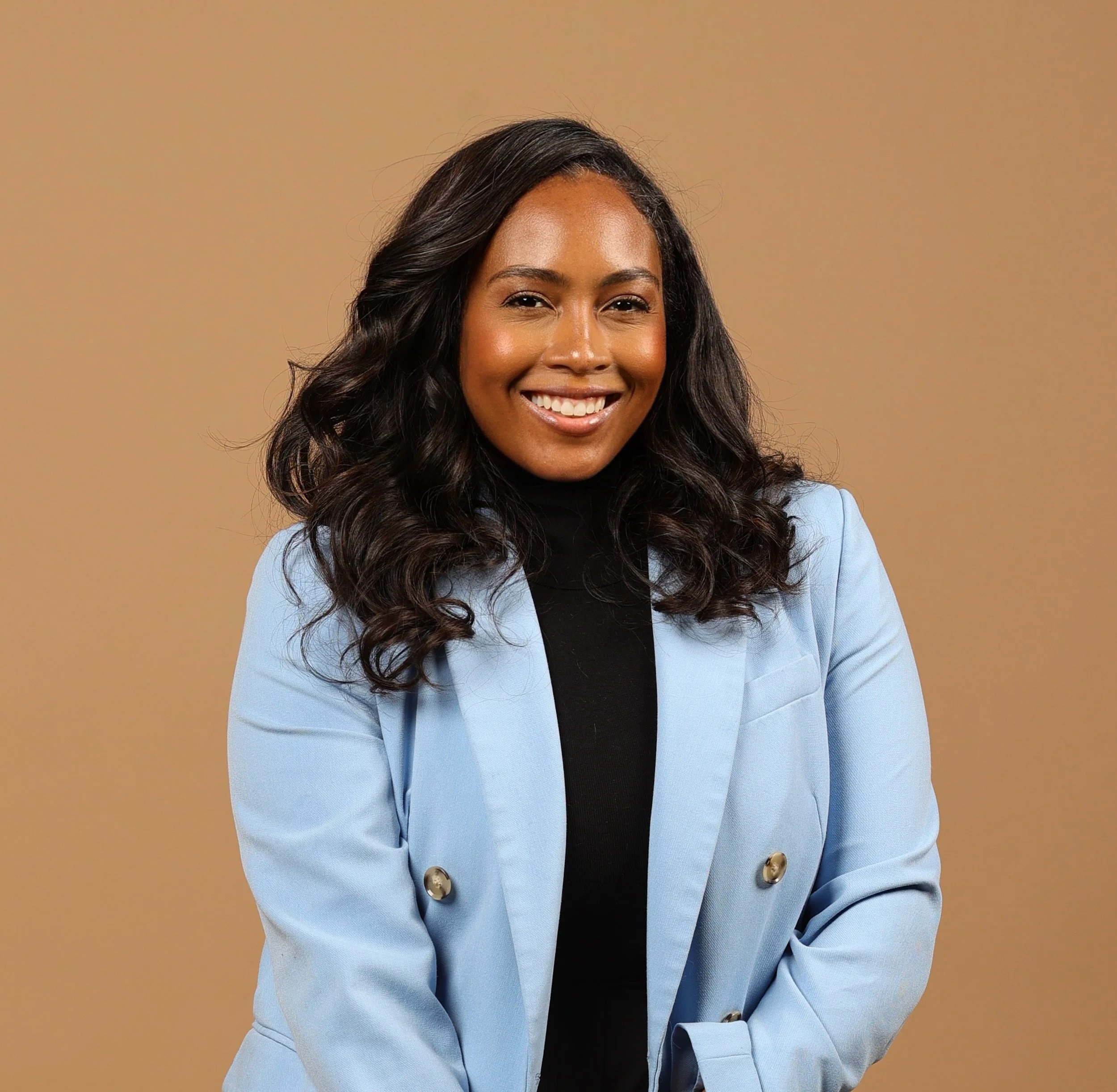 A professional portrait of California LMFT, Kalé Woods. A medium skin tone, african-american woman, long dark curly hair, wearing a black turtleneck and a light blue blazer, smiling against a beige background.