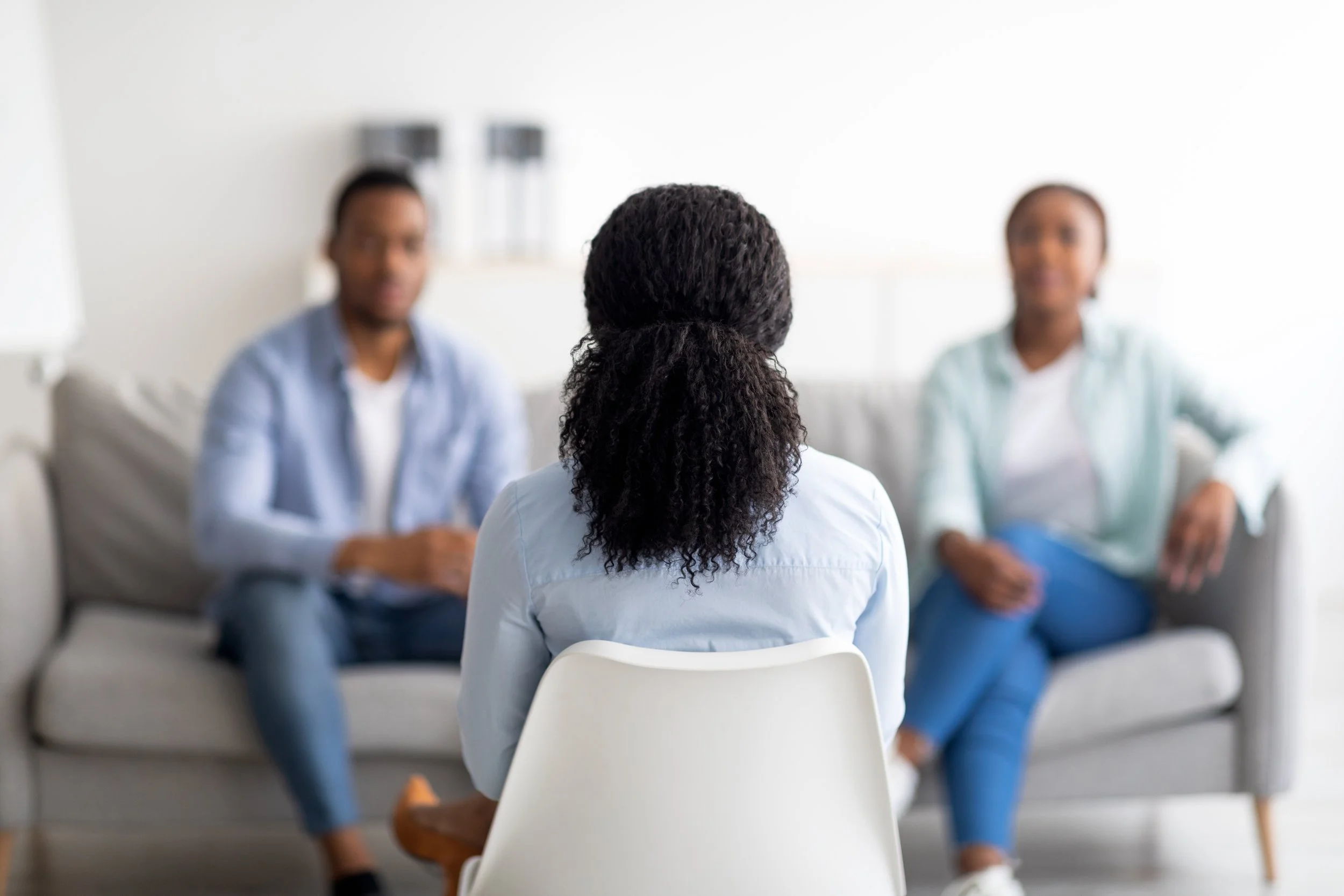 A woman sitting in an office or therapy room facing two people sitting on a sofa, both slightly blurred, while she listens attentively.
