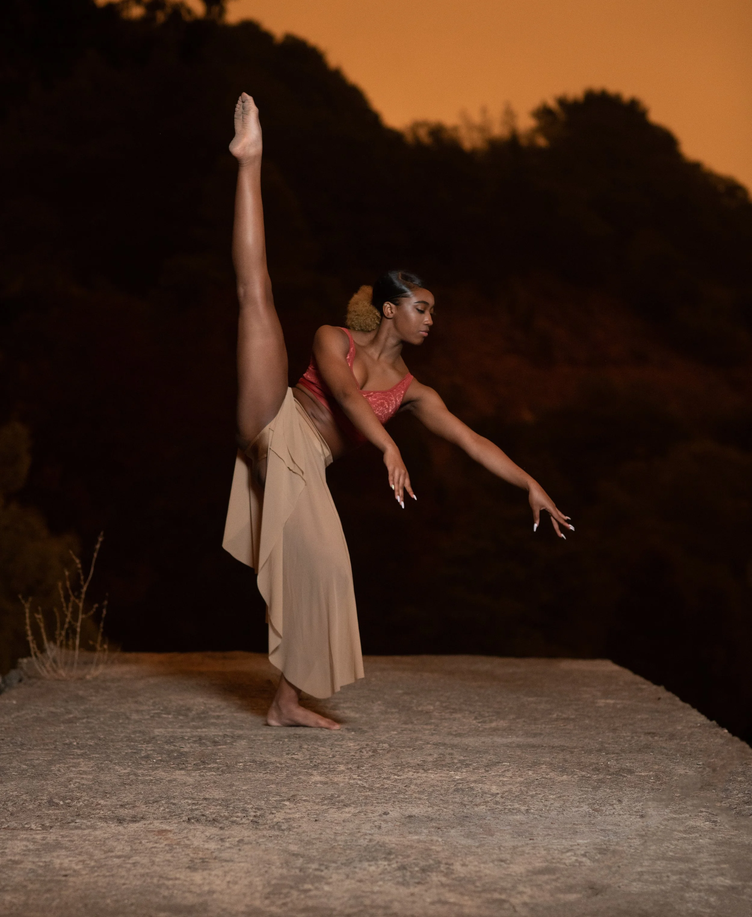 A woman in beige pants and a red top performing a high leg dance pose outdoors at sunset with a rocky terrain and trees in the background