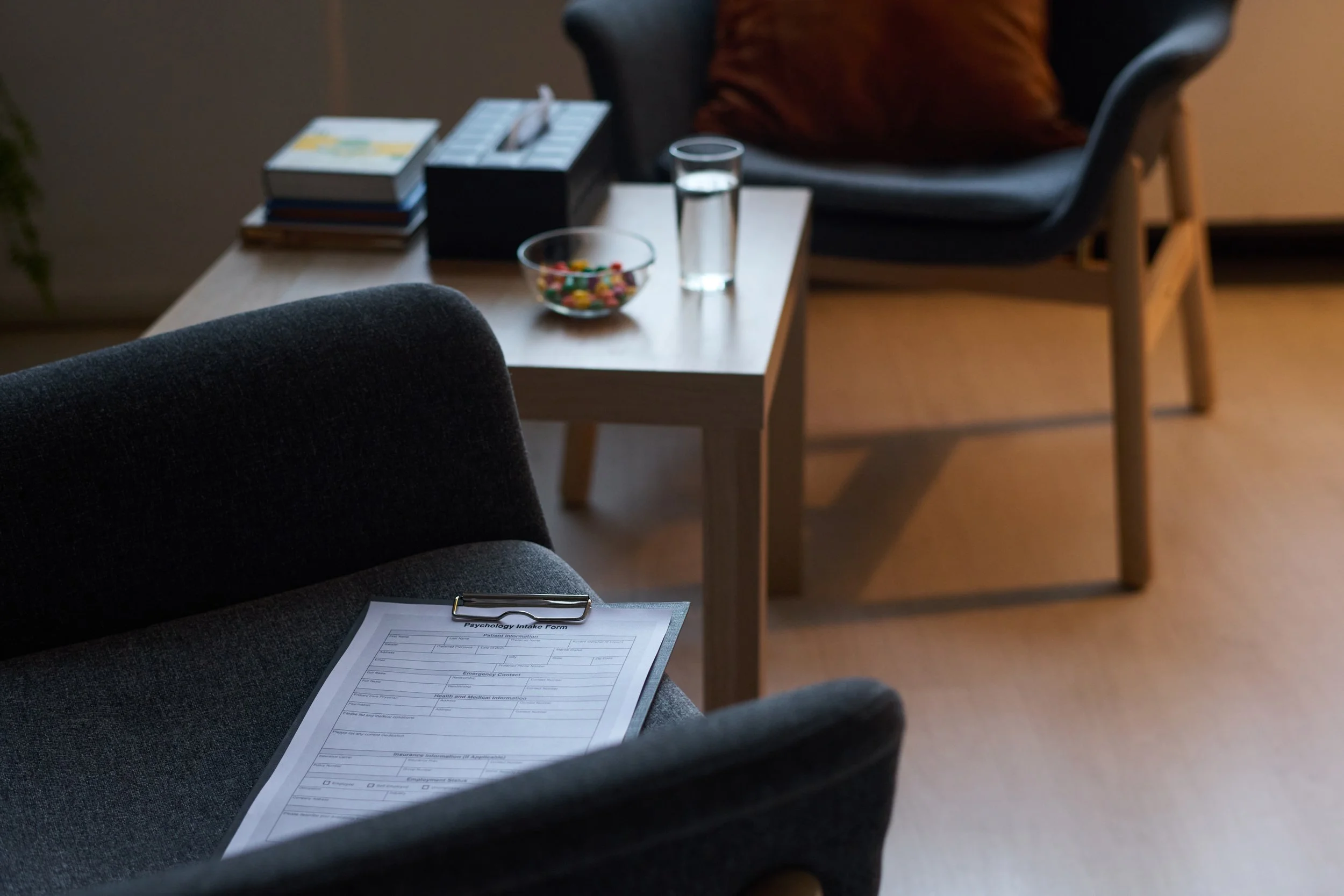 A waiting room with a clipboard on a dark gray armchair, a wooden table with a glass of water, a bowl of candies, a box of tissues, a stack of magazines, and two chairs with cushions in the background.