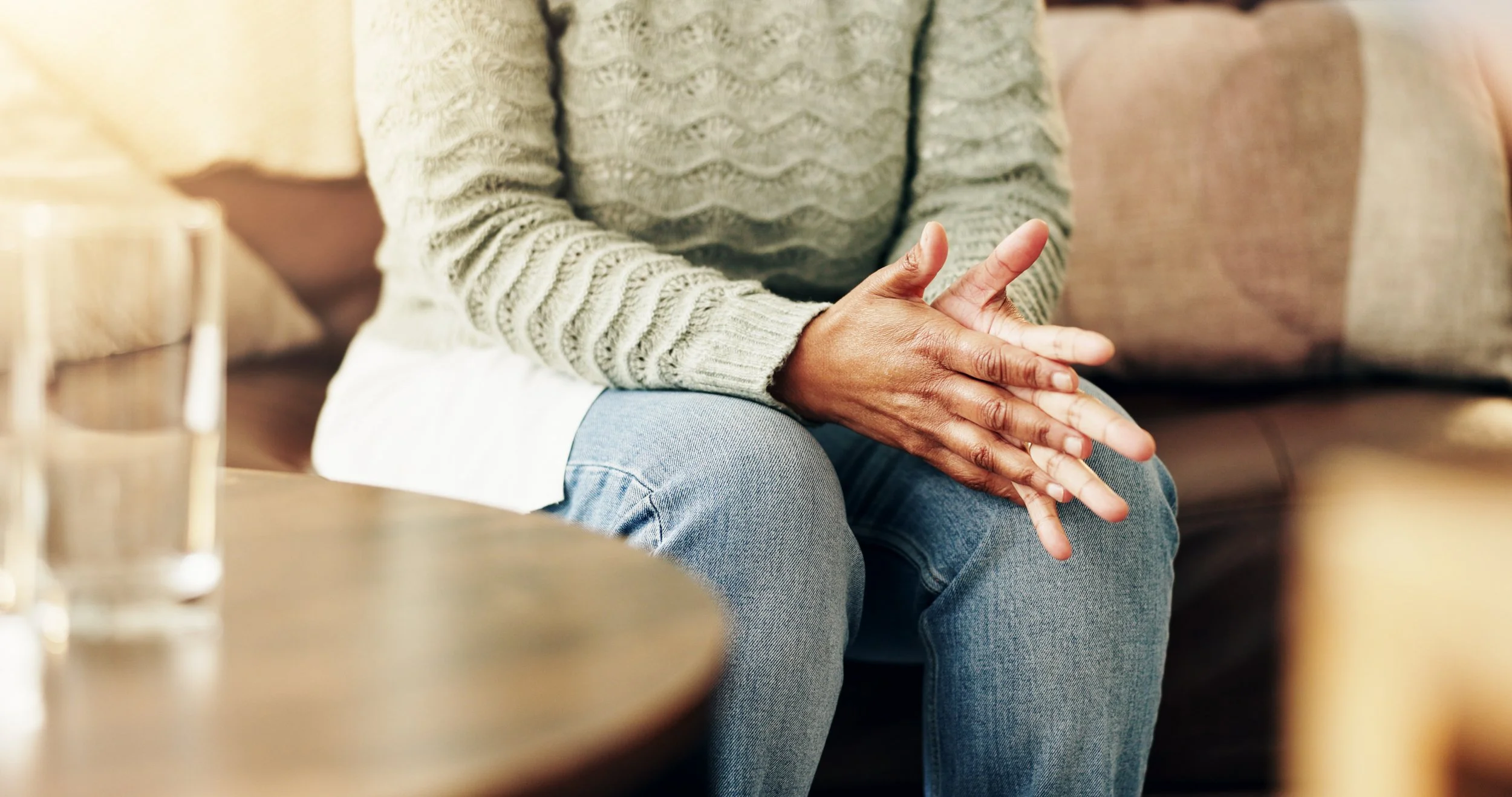 Person sitting on a sofa with clasped hands resting on their lap, wearing a beige sweater and blue jeans, in a cozy living room.