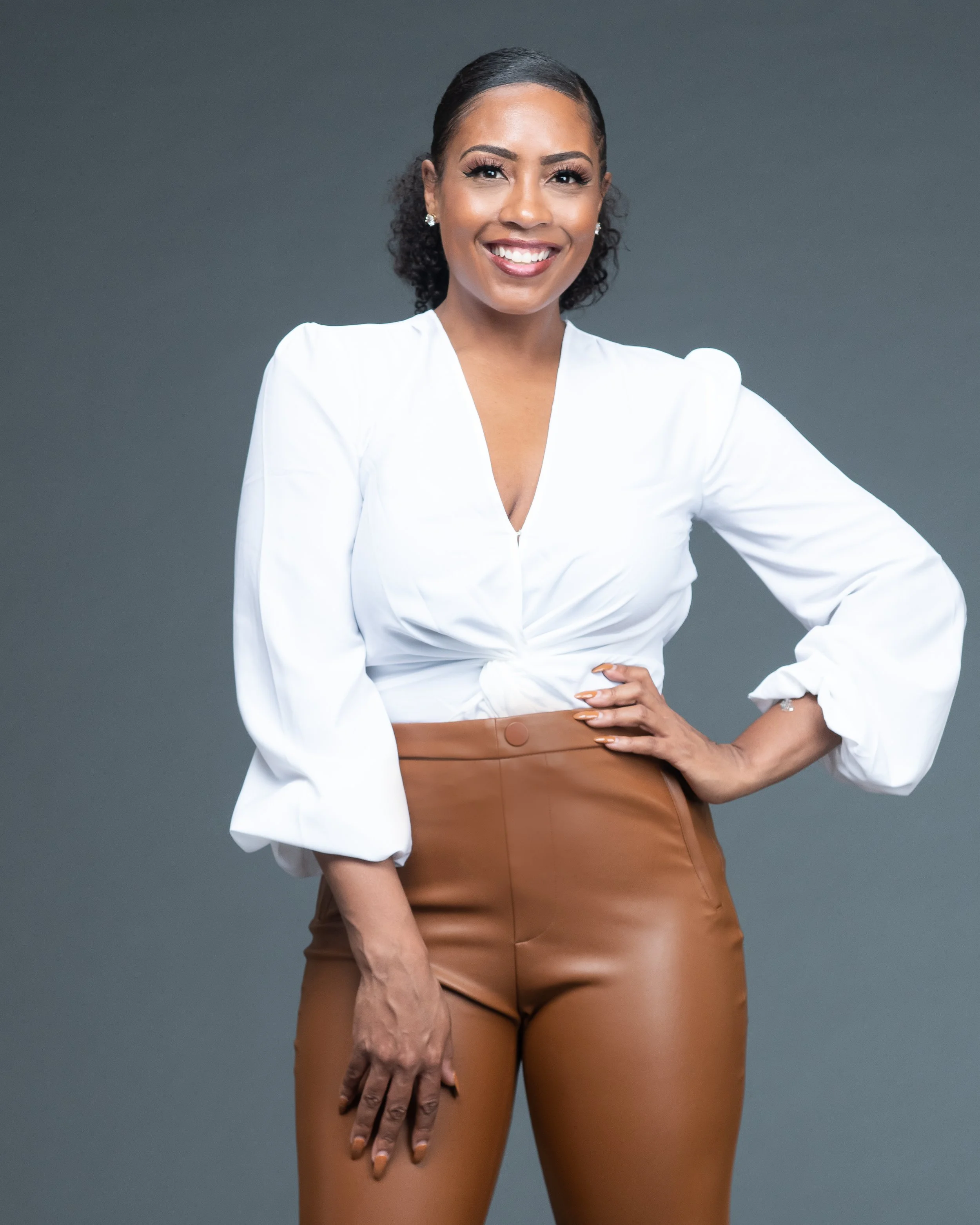 A woman with dark hair, wearing a white blouse and brown pants, smiling with her hand on her hip against a gray background.