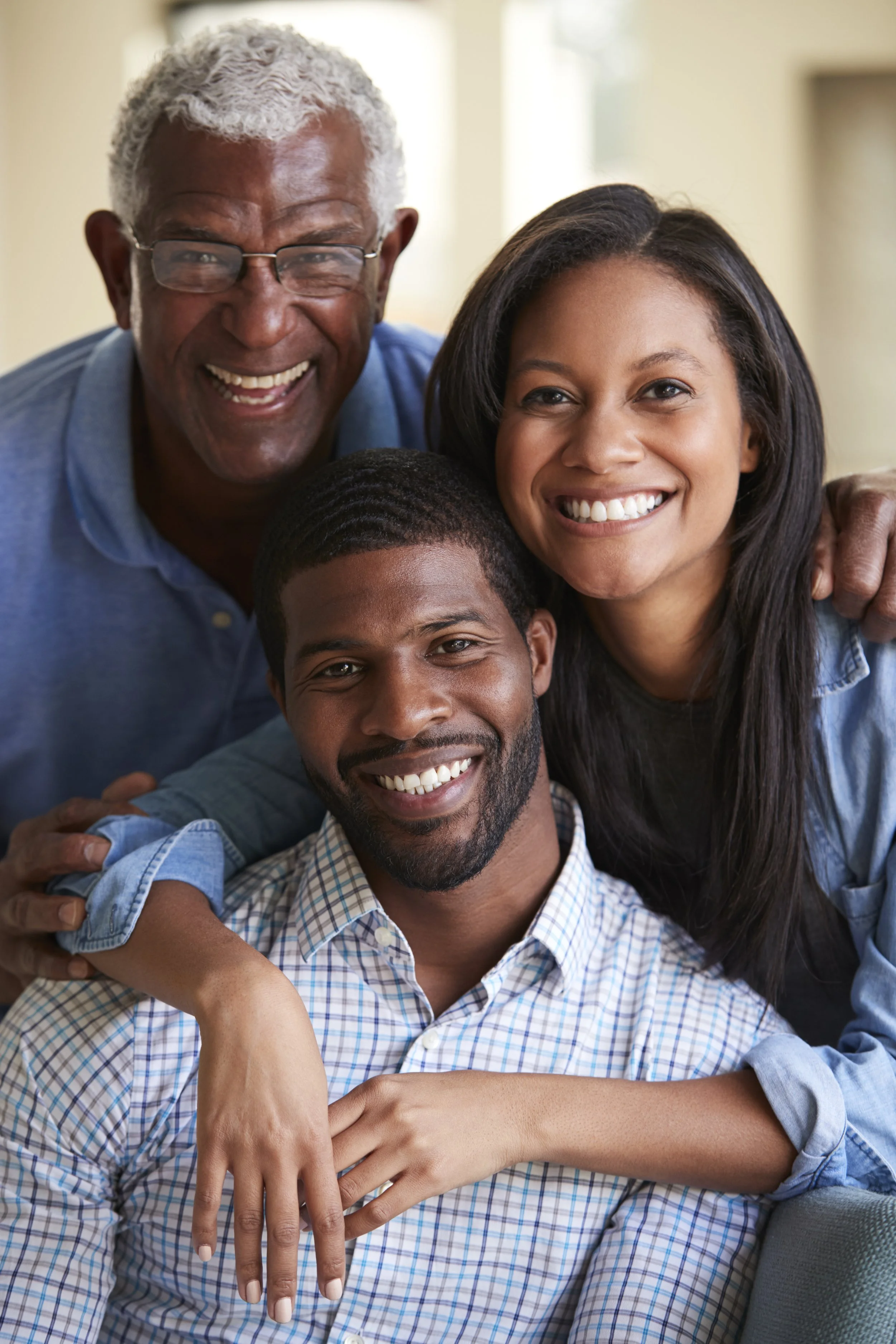 A diverse family group: an older man, a woman, and a younger man, all smiling and hugging in a bright indoor setting.