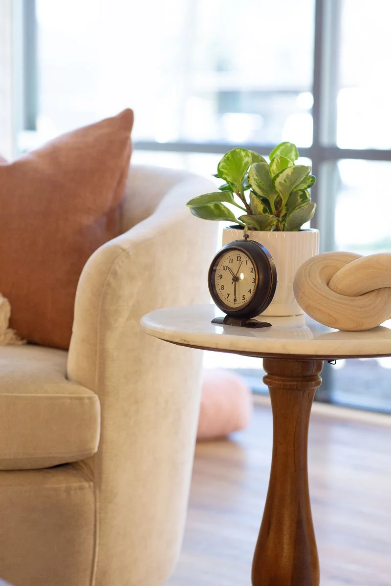 Close-up of a side table with a potted plant, a small clock, and a decorative wooden sculpture in a bright, cozy living room.