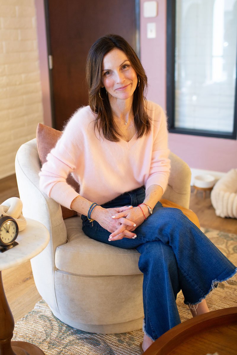 Rebecca Tofield sitting on a beige armchair in cozy office, wearing a light pink sweater and blue jeans, smiling at the camera.