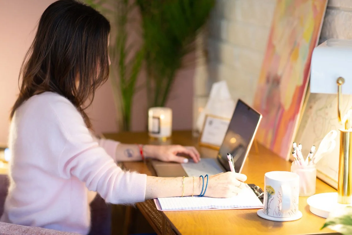 Rebecca Tofield sitting at her desk writing in a notebook with a pen, in a cozy, warmly-lit room.