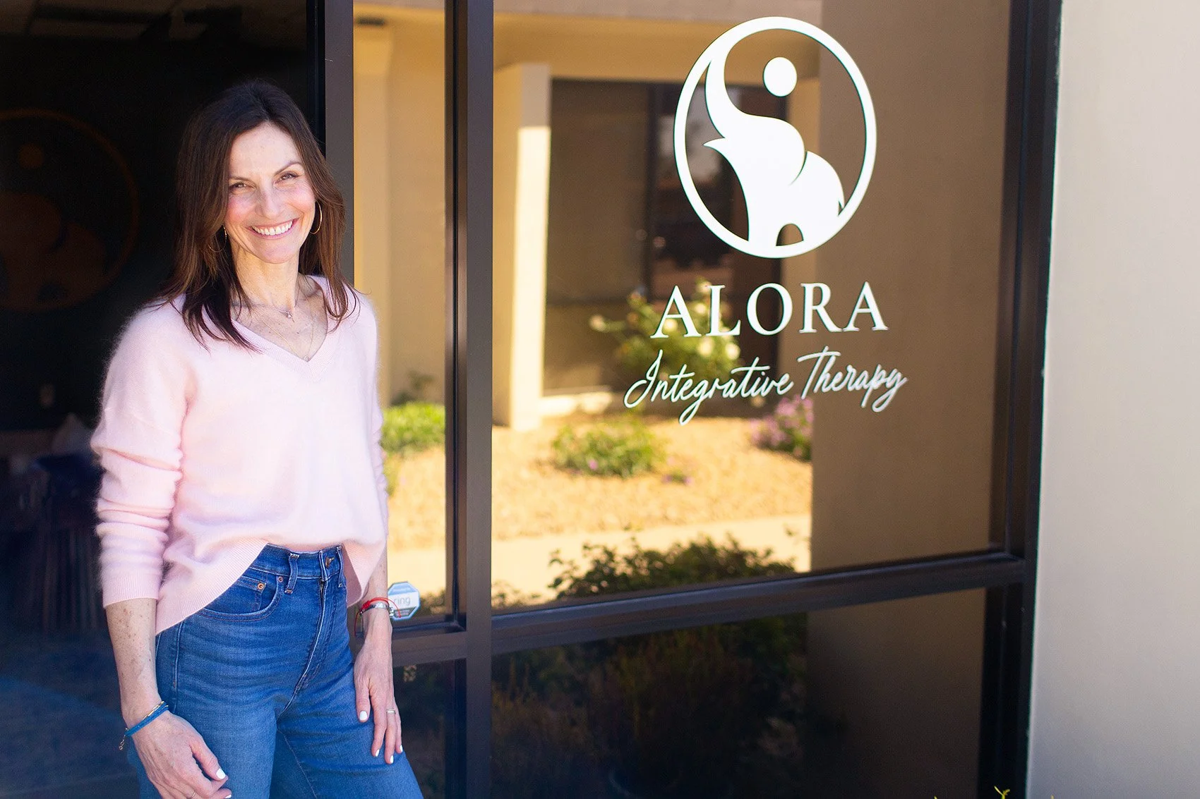 Rebecca standing outside Alora Integrative Therapy in front of glass door with her logo on it.
