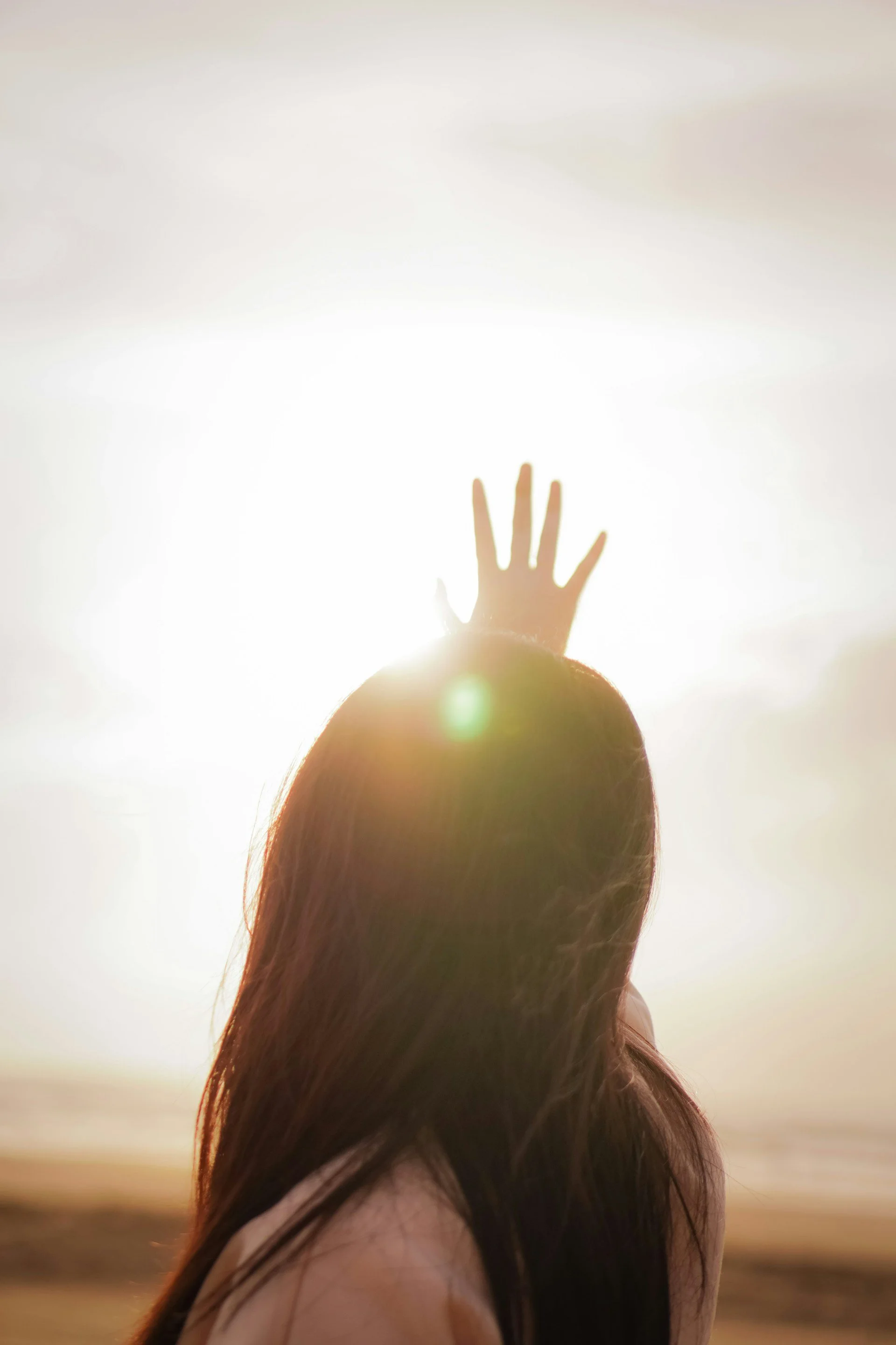 A person with long hair reaching towards the sky with their hand, standing on a beach during sunset, with the sun shining brightly behind them.