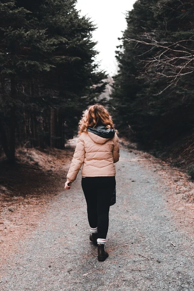 A woman walking on a dirt trail through a forest of tall coniferous trees, wearing a pale pink puffer jacket.