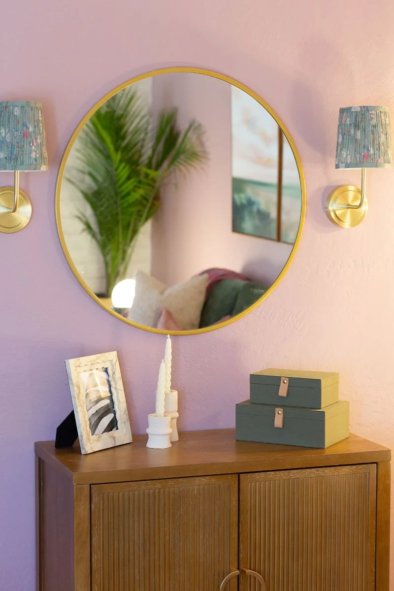 A round mirror, two wall-mounted blue lamps, a wooden cabinet topped with picture frame, candles, and decorative boxes.
