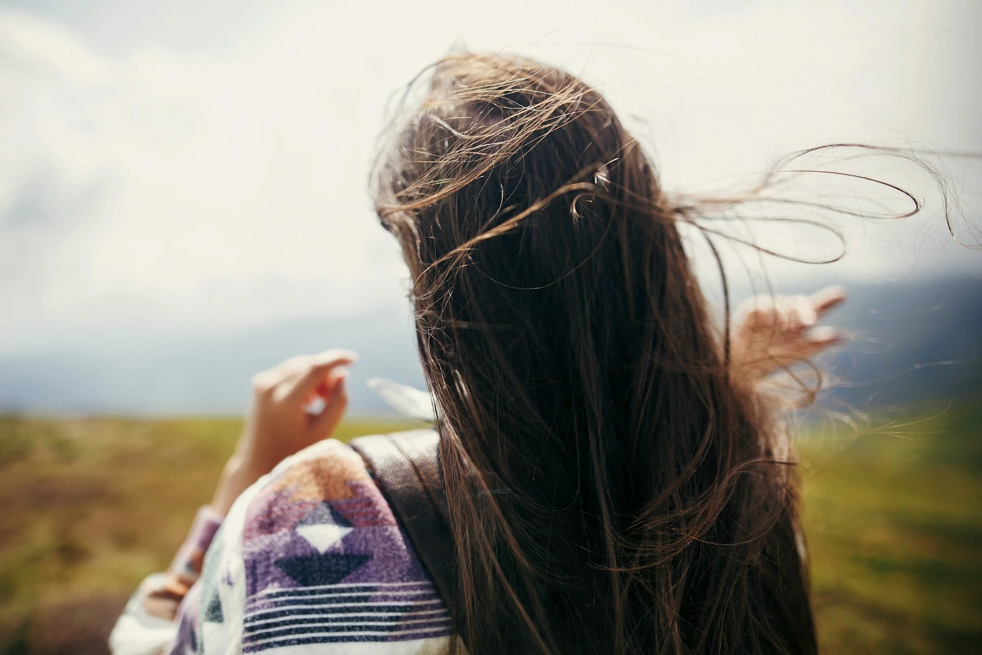 A woman with long hair standing outdoors with wind blowing through her hair, wearing a patterned sweater.