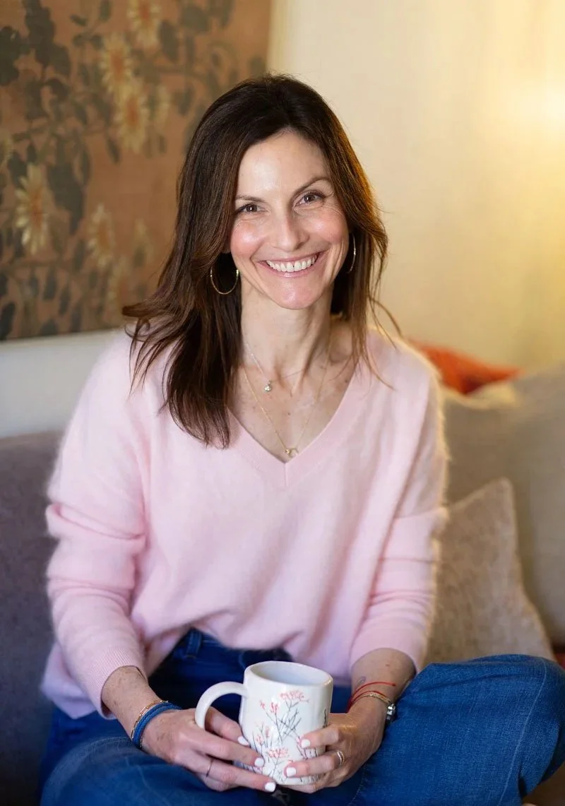 Rebecca Tofield, sitting on a sofa, holding a white mug with floral design, wearing a light pink sweater in a cozy room therapy office in AZ.