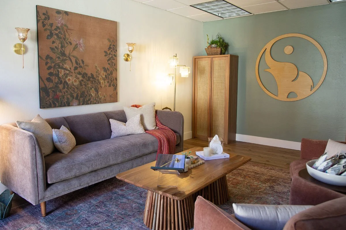 Waiting area with a soft gray sofa, a wooden coffee table, wall art, and the Alora Integrative Therapy elephant logo.