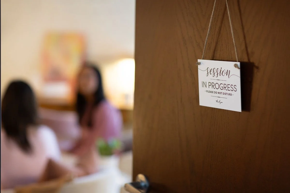 A wooden door with a sign that reads 'session in progress - please do not disturb', with blurred people in the background sitting at a table.