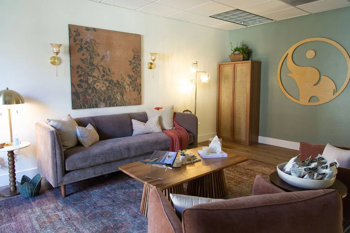 Therapist waiting room with a gray sofa, throw pillows, a wooden coffee table, a standing lamp, wall-mounted lamps, a large wall artwork, a wooden cabinet, and decorative items.