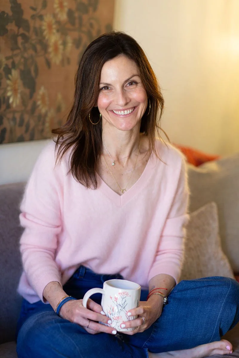 Rebecca Tofield wearing a light pink sweater, smiling while sitting on a sofa and holding a white mug with a floral design, in a warmly lit room.