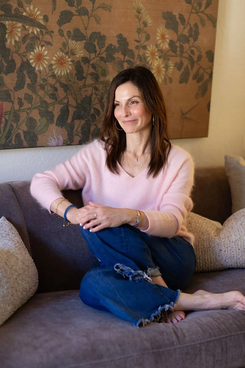 Rebecca Tofield sitting on a sofa with a floral painting on the wall behind her, wearing a pink sweater and blue jeans, smiling gently.