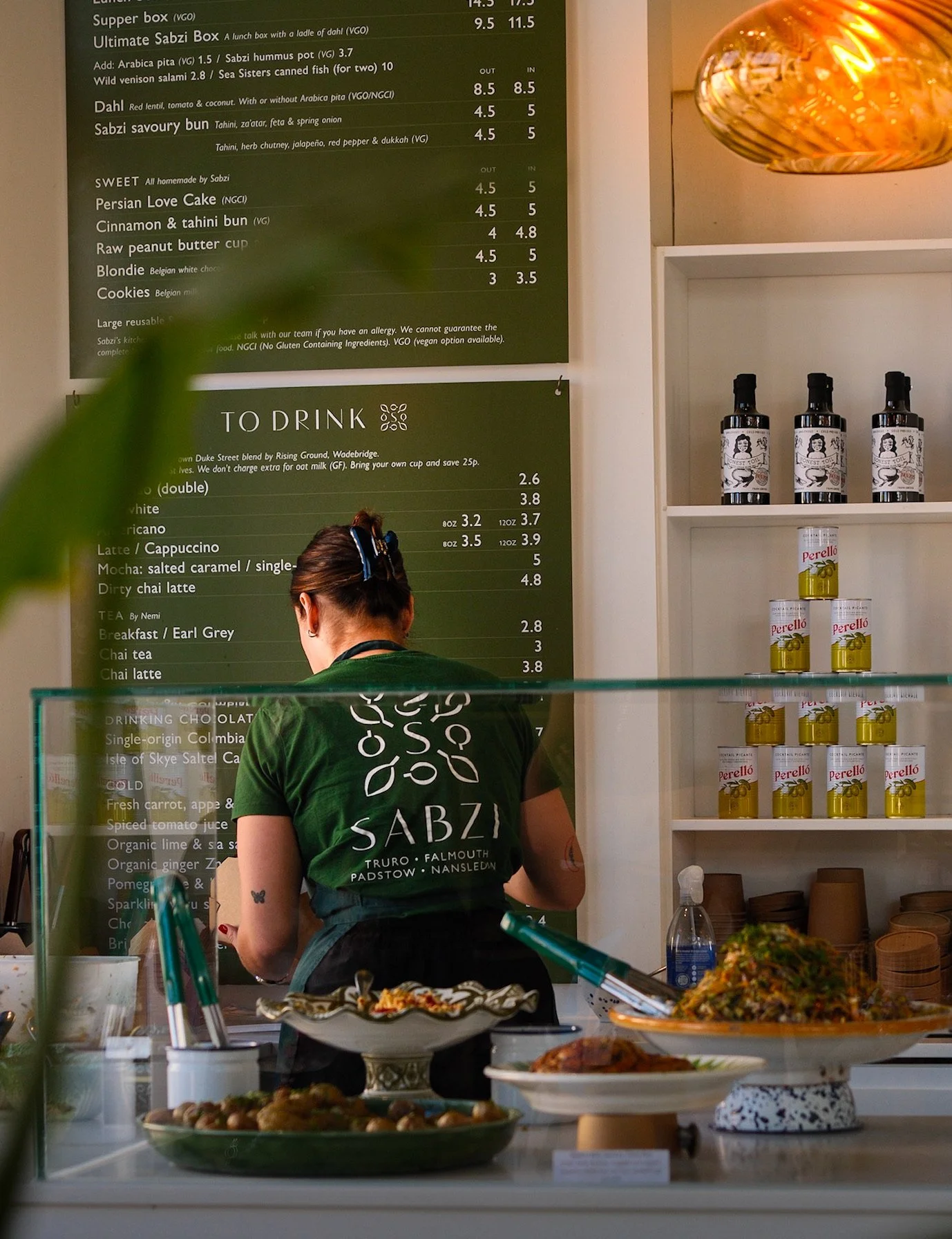A person in a green Sabzi apron preparing food behind a glass display counter, with plates of food in front and a green menu board on the wall behind them in a restaurant or cafe.