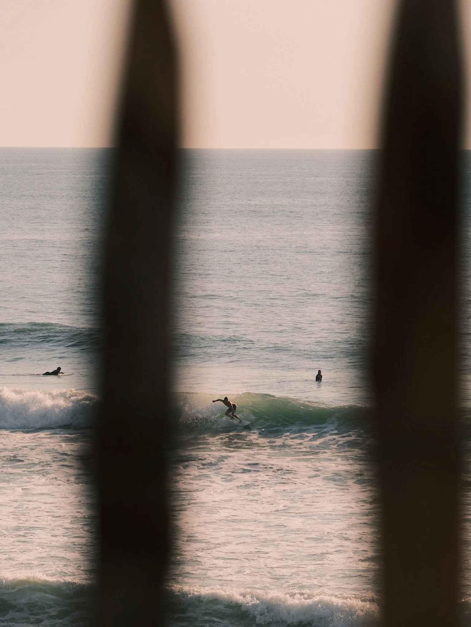 View of an ocean with gentle waves during sunset, viewed through black railings, with a surfer riding a wave and other people in the water.
