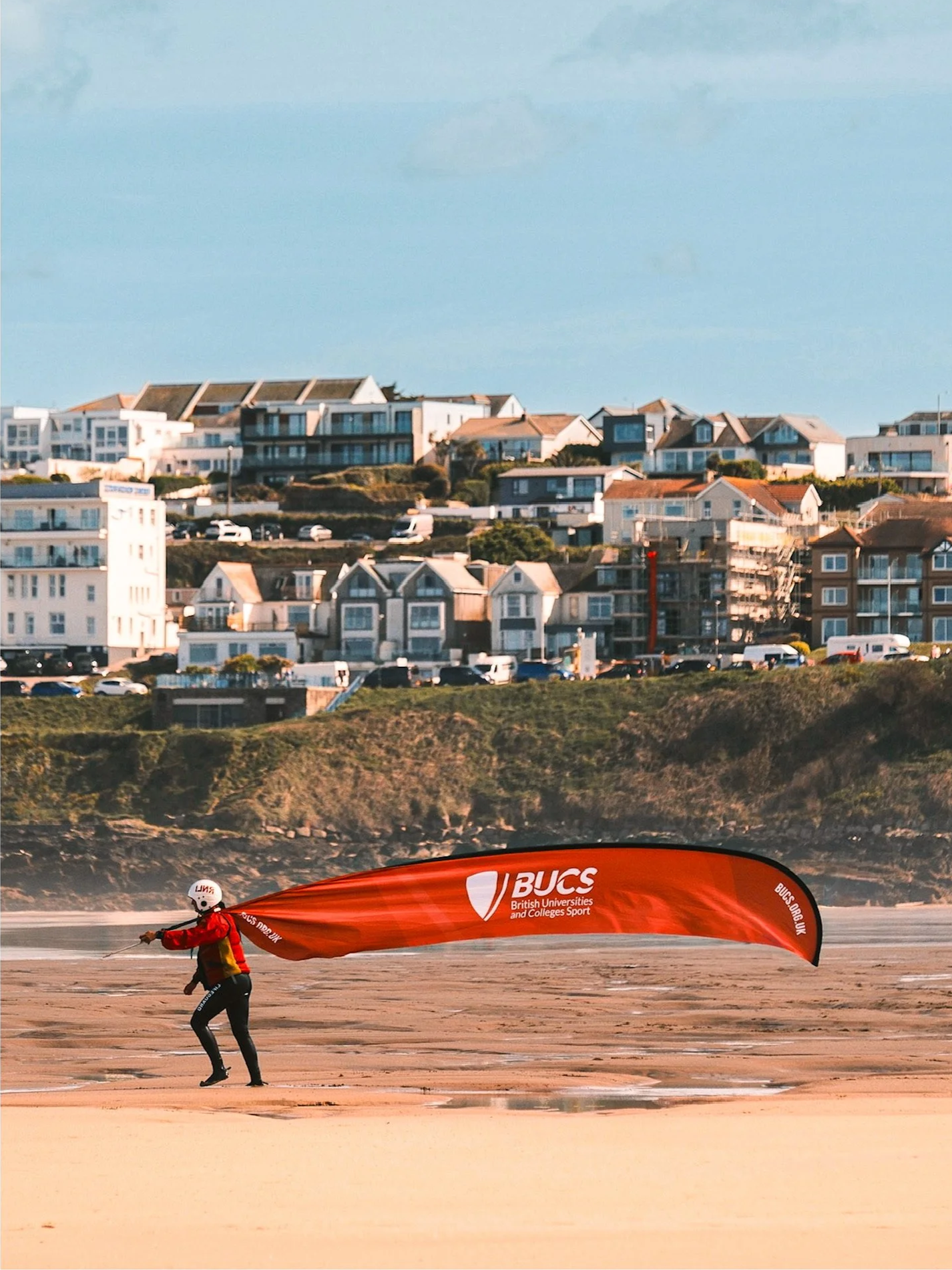 Person on the beach holding a large orange banner with the logo of British Universities and Colleges Sport (BUCS).