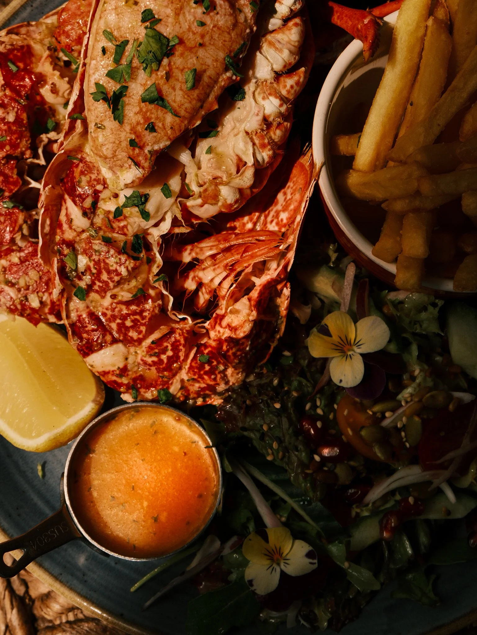 A seafood platter featuring lobster tail garnished with chopped herbs, a cup of orange-colored sauce, a lemon wedge, and a side of French fries. The platter also includes a salad with edible flowers and mixed greens.