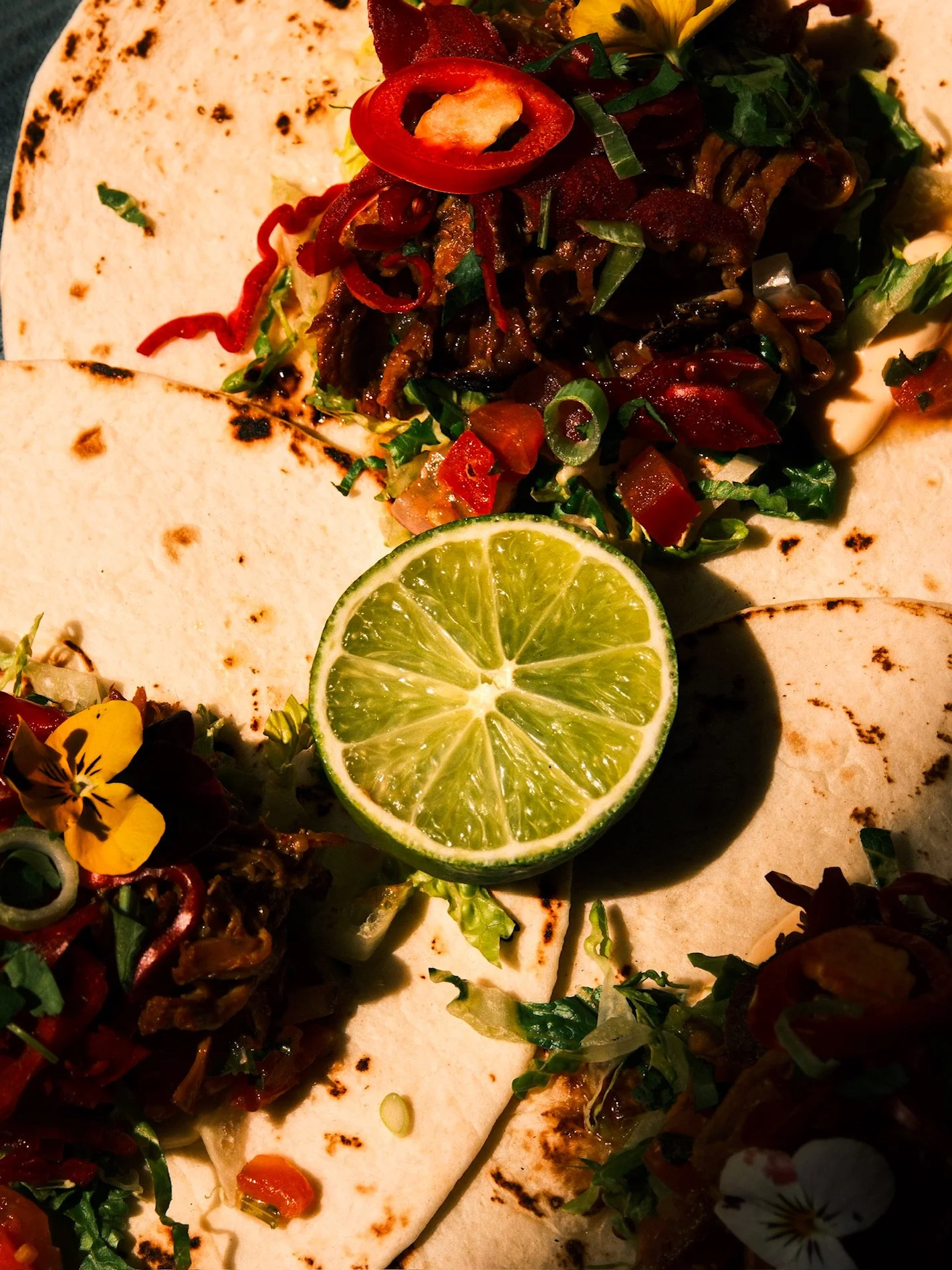 Close-up of a taco with grilled meat, chopped vegetables, and sliced red chili peppers, garnished with a lime wedge and edible flowers.