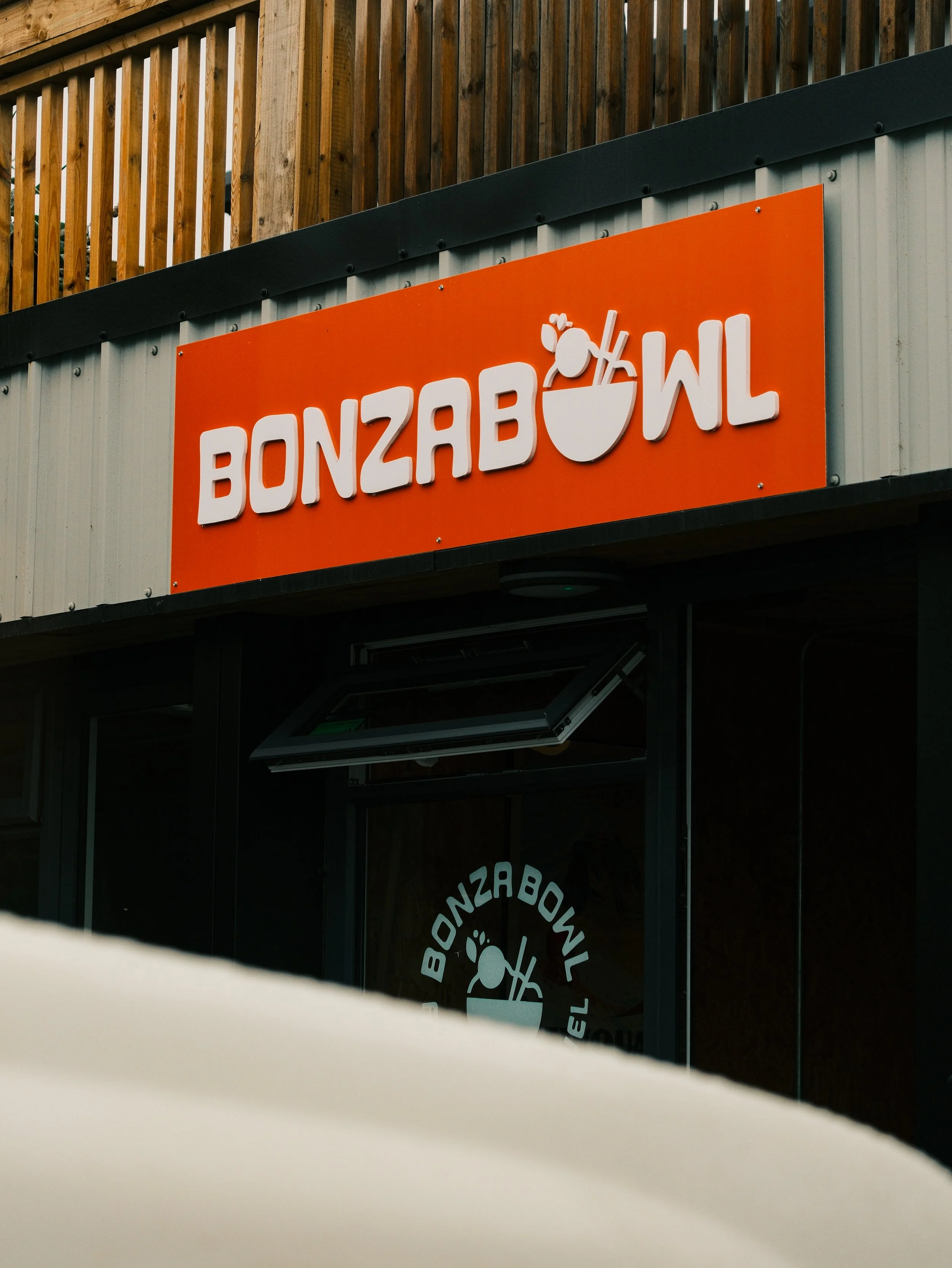 Close-up of a sign for Bonzabowl, with part of the reflection in a window showing the logo and some wooden fencing above.