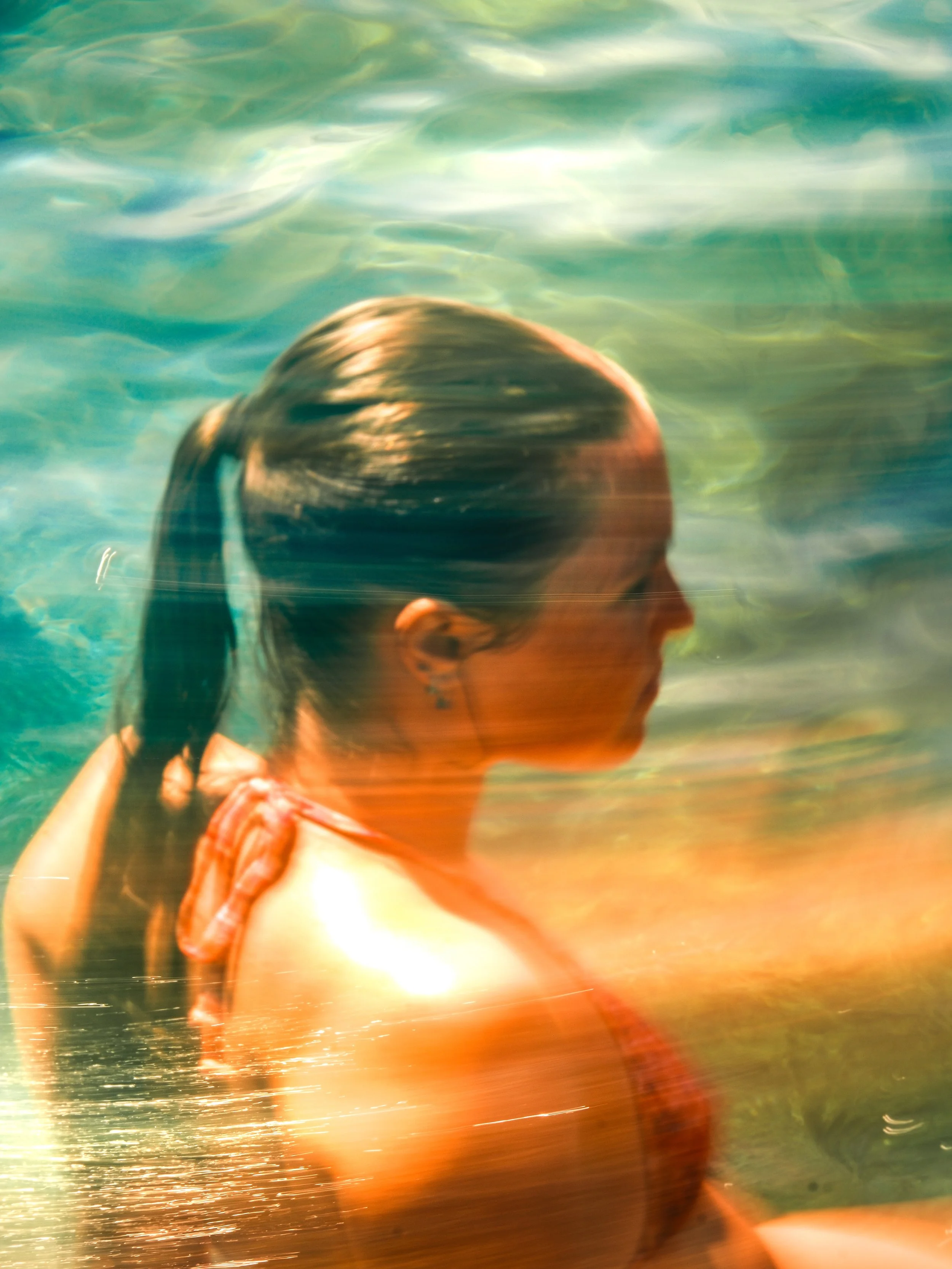 A person swimming in a body of water, with a close-up of their head and arm visible, and sunlight reflecting on the surface.